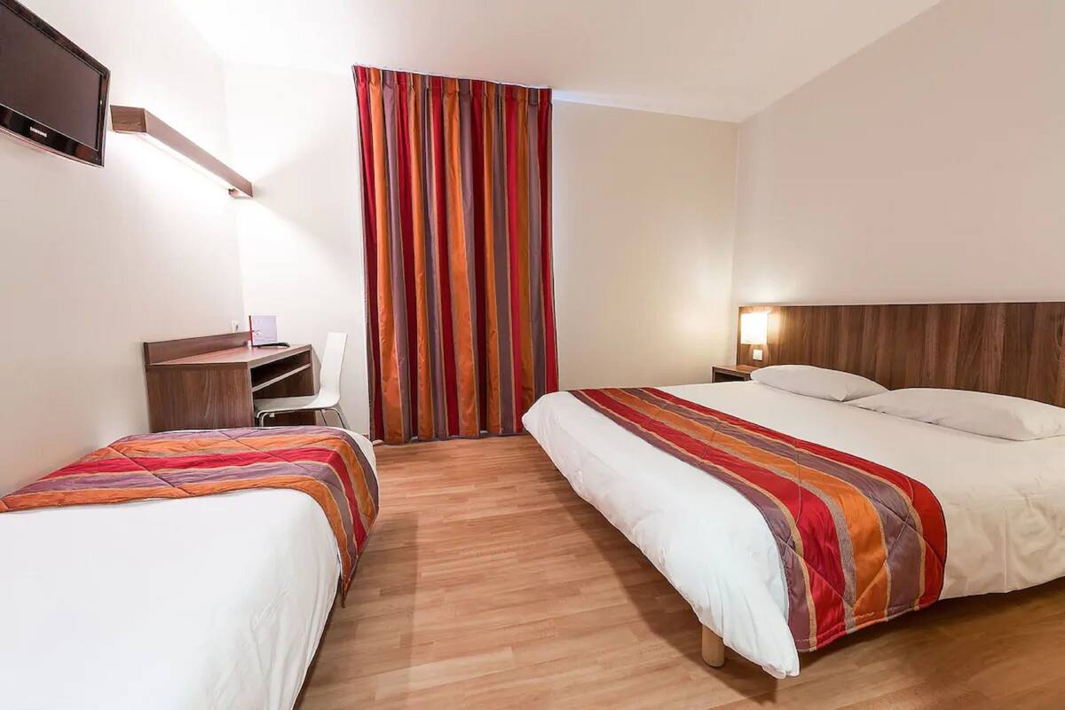 Quarto de hotel com duas camas, uma escrivaninha, televisão e janela com cortinas listradas.