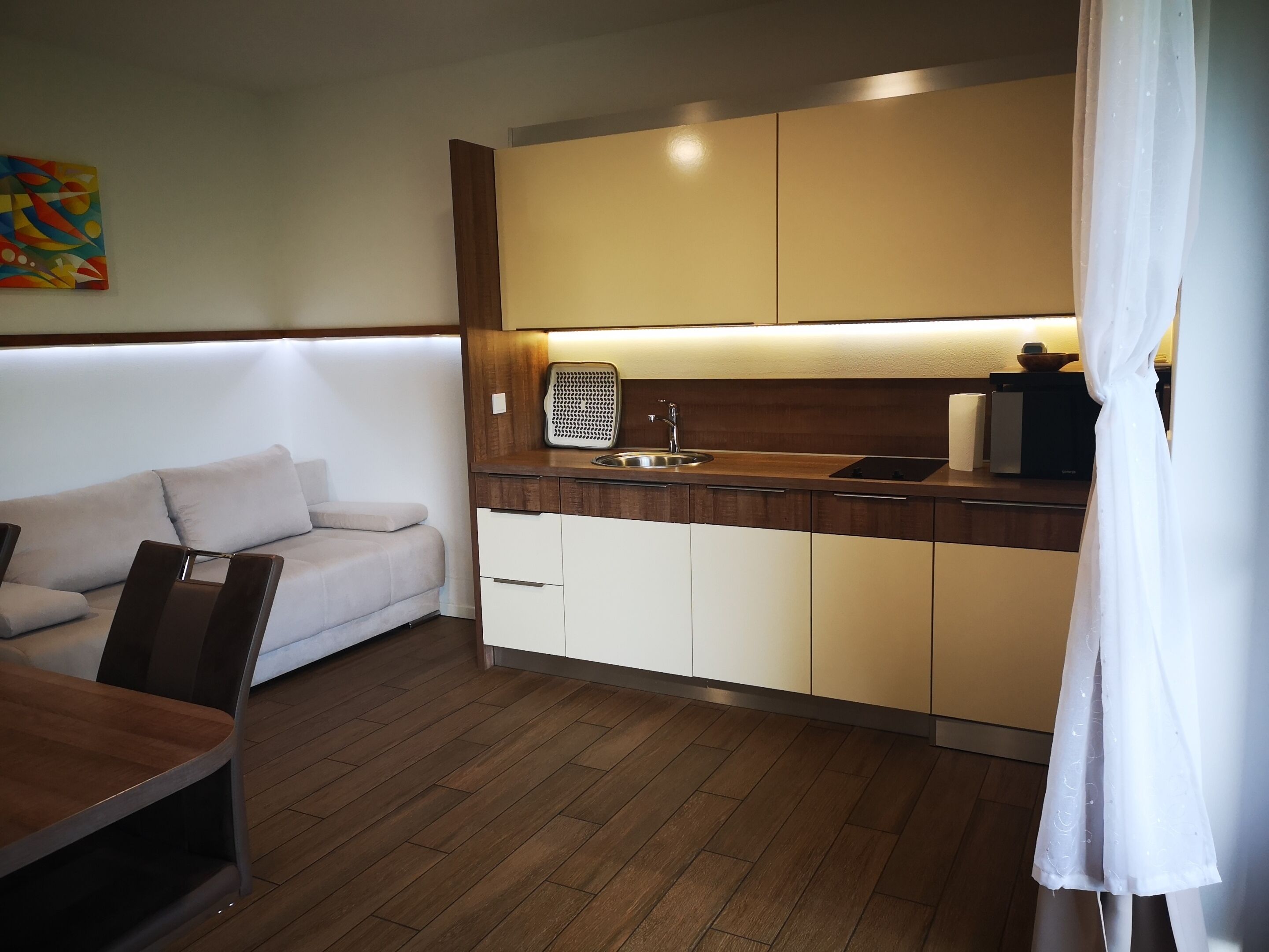 A compact kitchenette with a sink, countertop, and wooden cabinets. A sofa and a table are visible in the adjacent living area.