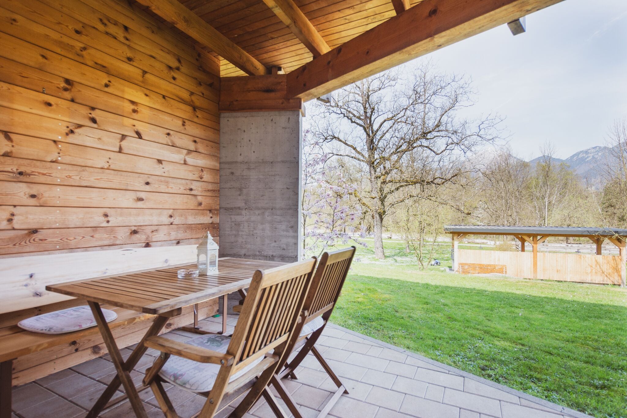 Una terraza de madera con una mesa y sillas, un muro de hormigón y vistas a un parque con árboles y montañas.