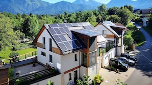 A modern house with solar panels on the roof, surrounded by lush greenery and mountains in the background.