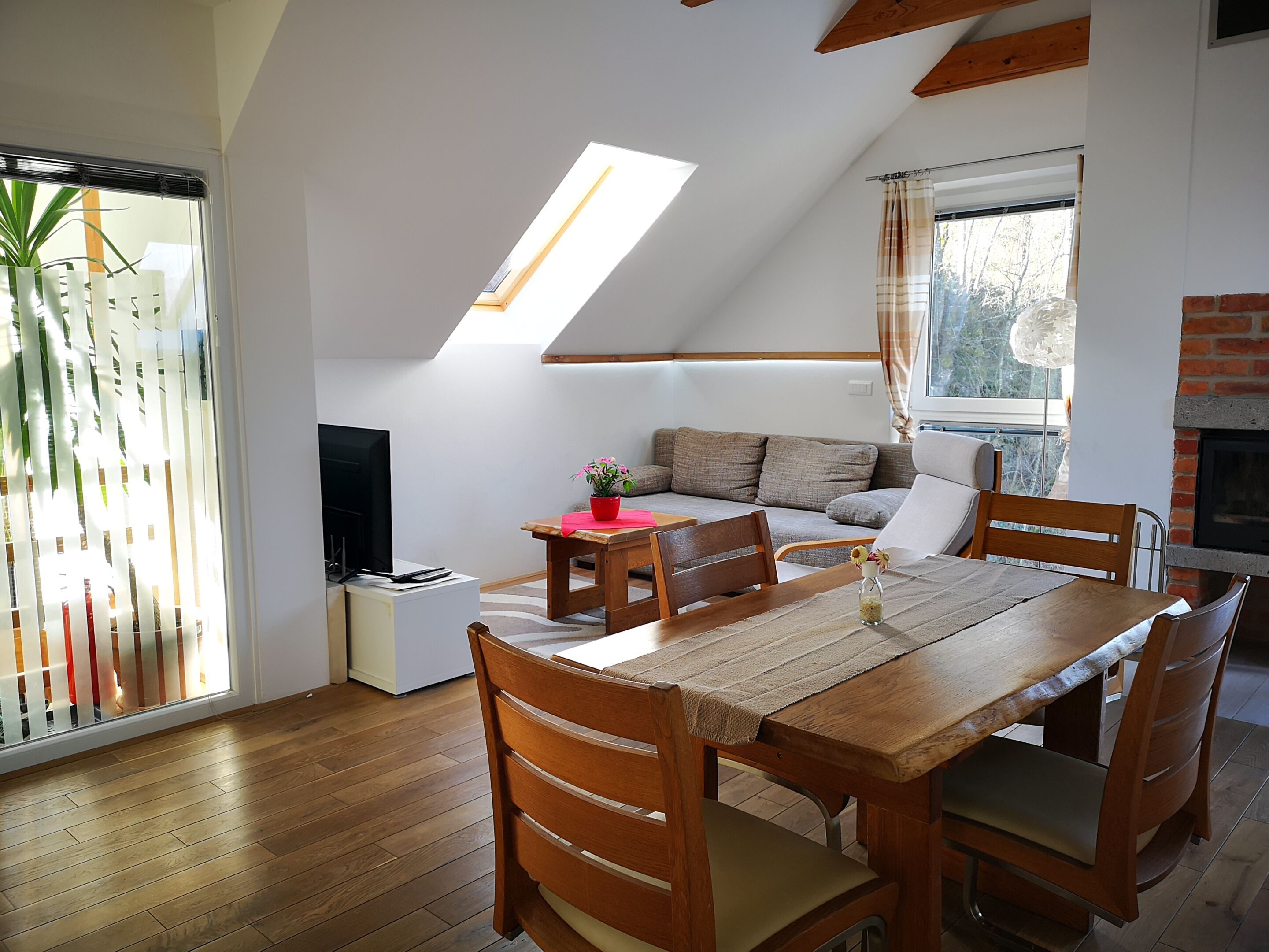 Una habitación acogedora con una mesa de comedor de madera, sillas, un sofá y un televisor. Hay una ventana con cortinas y una planta, además de una chimenea.