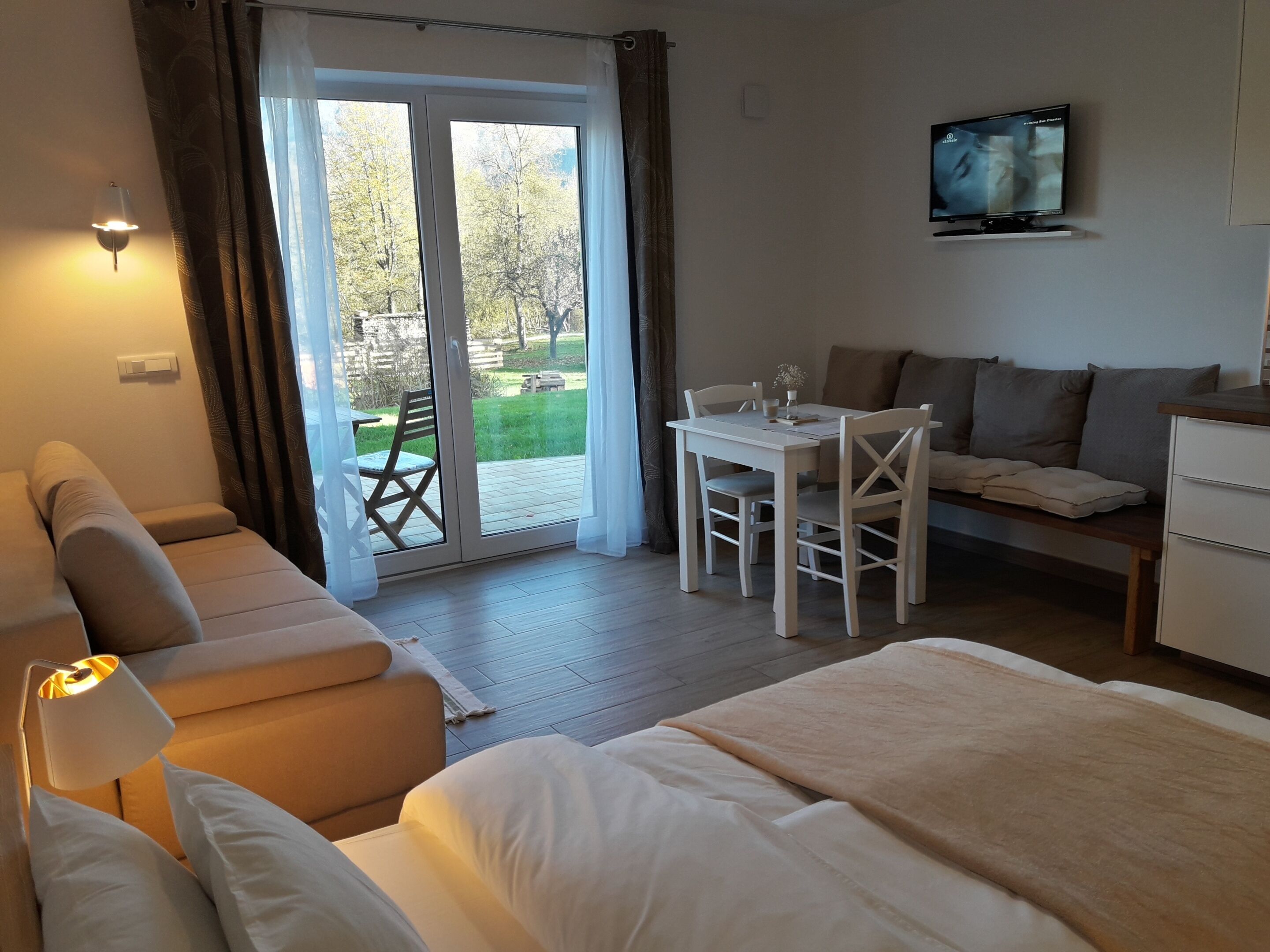 A hotel room with a bed, a sofa, a dining table with chairs, a TV, and a sliding glass door leading to a garden.