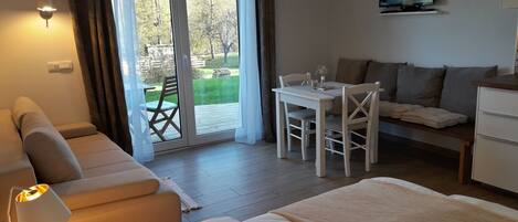 A hotel room with a bed, a sofa, a dining table with chairs, a TV, and a sliding glass door leading to a garden.