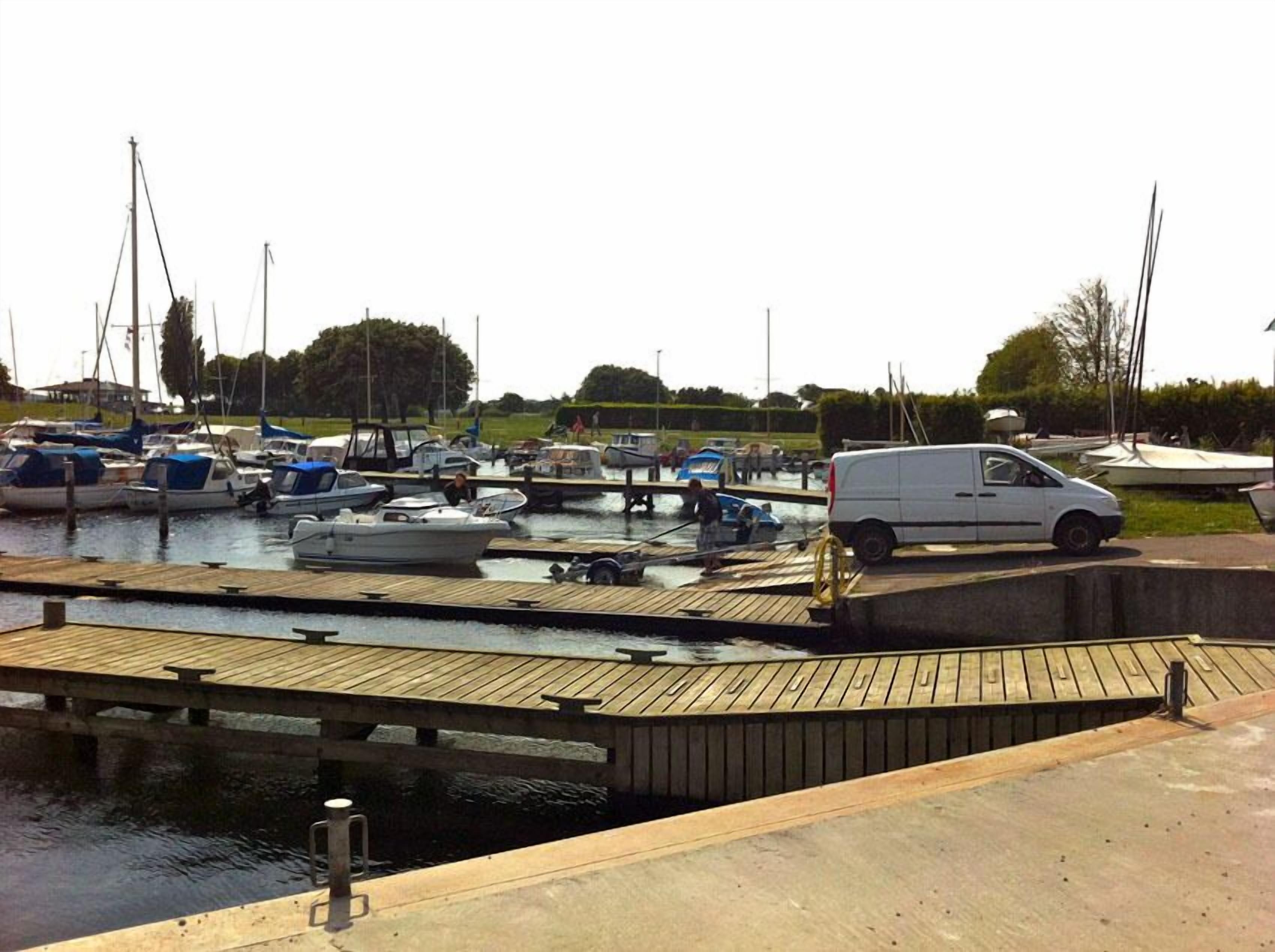 Un port de plaisance avec des bateaux amarrés, un fourgon blanc garé sur une jetée en bois et une promenade en béton menant à l’eau.
