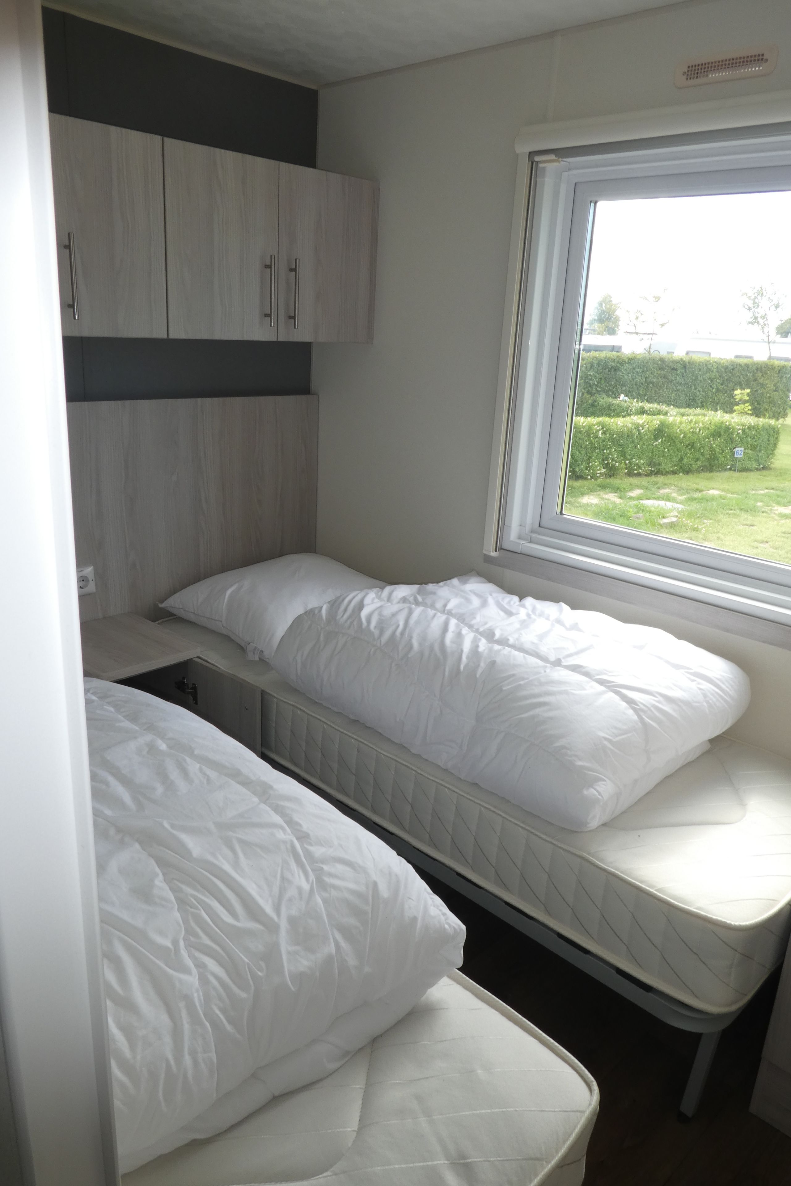 A compact bedroom with two beds, a window with a view of greenery, and built-in cabinets.