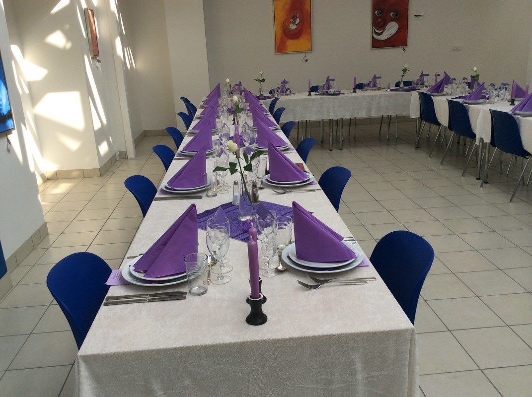 Ruokailutila, jossa on katetut pöydät valmiina ateriaa varten, ja koristeena violetit servetit sekä lasiesineet.