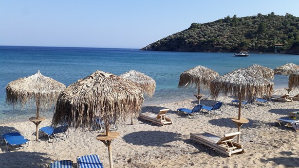 A beach with thatched umbrellas and sun loungers, a clear blue sea, and a small island in the background. - Meltemia Studios Samos (Samos)