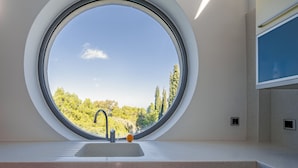 A round window with a sink and faucet, a blue wall, and a view of trees and sky. - Minimal Blue Villa (Markopoulo Mesogaias)