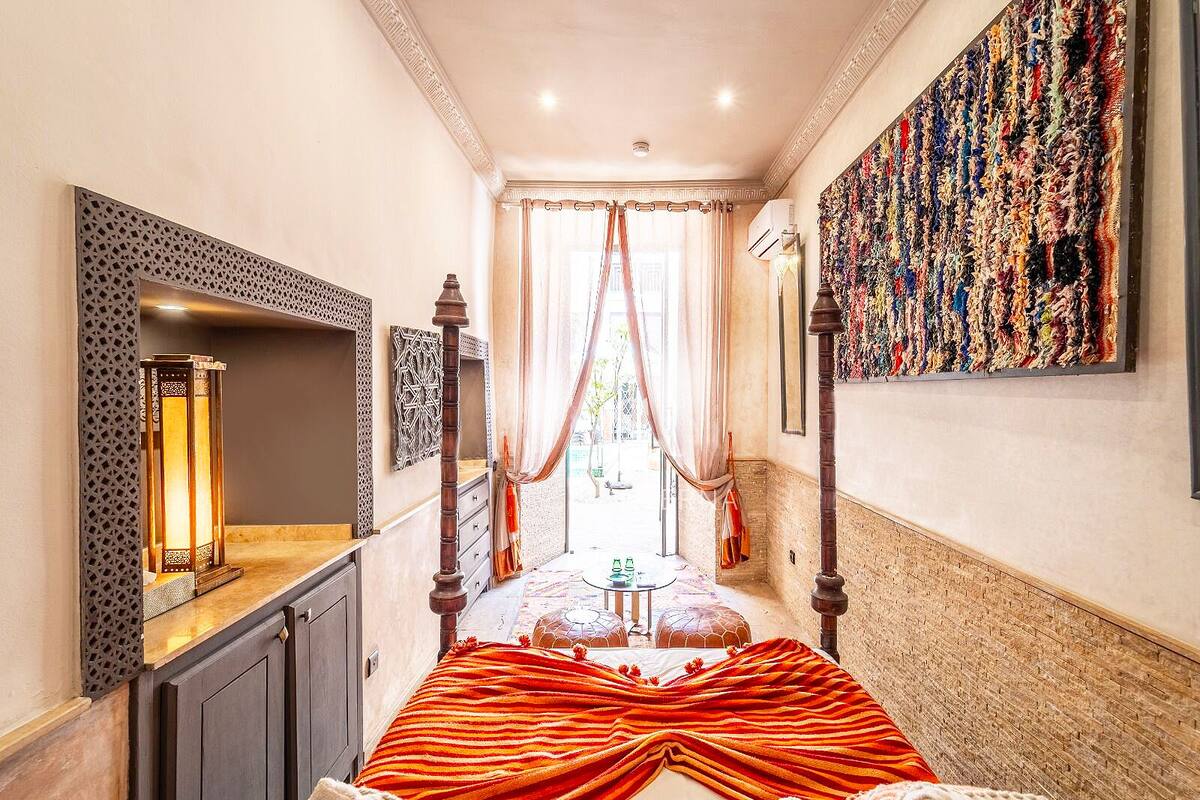 A bedroom with a four-poster bed, a large window with curtains, a decorative wall hanging, and a small table with a rug.