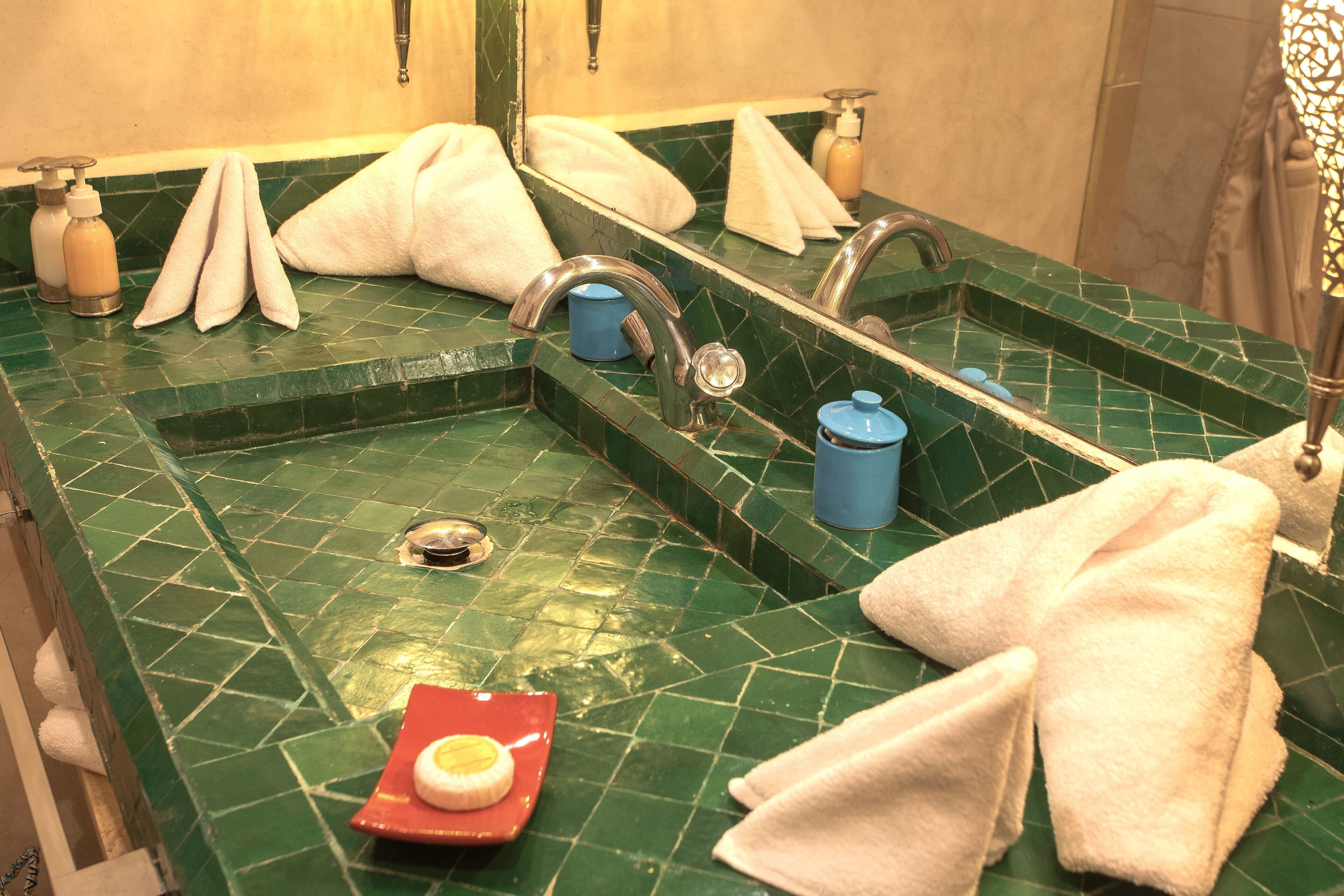 A bathroom with a green tiled sink, two faucets, and a mirror. There are towels and toiletries on the counter.