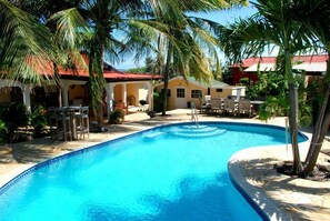 A swimming pool with a tiled edge, surrounded by palm trees and a building with a red roof.