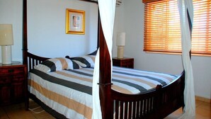A four-poster bed with a striped bedspread, a bedside table with a lamp, and a window with blinds. - Paradera Paradise (Paradera)