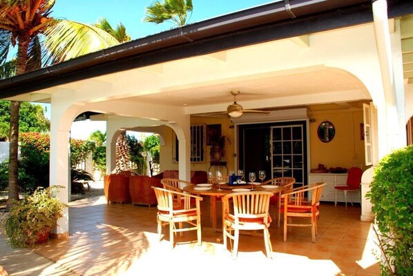 A covered outdoor dining area with a table and chairs, surrounded by plants and a view of the house.