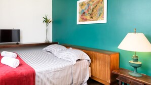 Quarto de hotel com uma cama, televisão, escrivaninha com luminária e um quadro na parede.