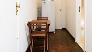 Um corredor estreito com uma mesa de madeira e cadeiras, uma porta branca e uma geladeira branca ao fundo.