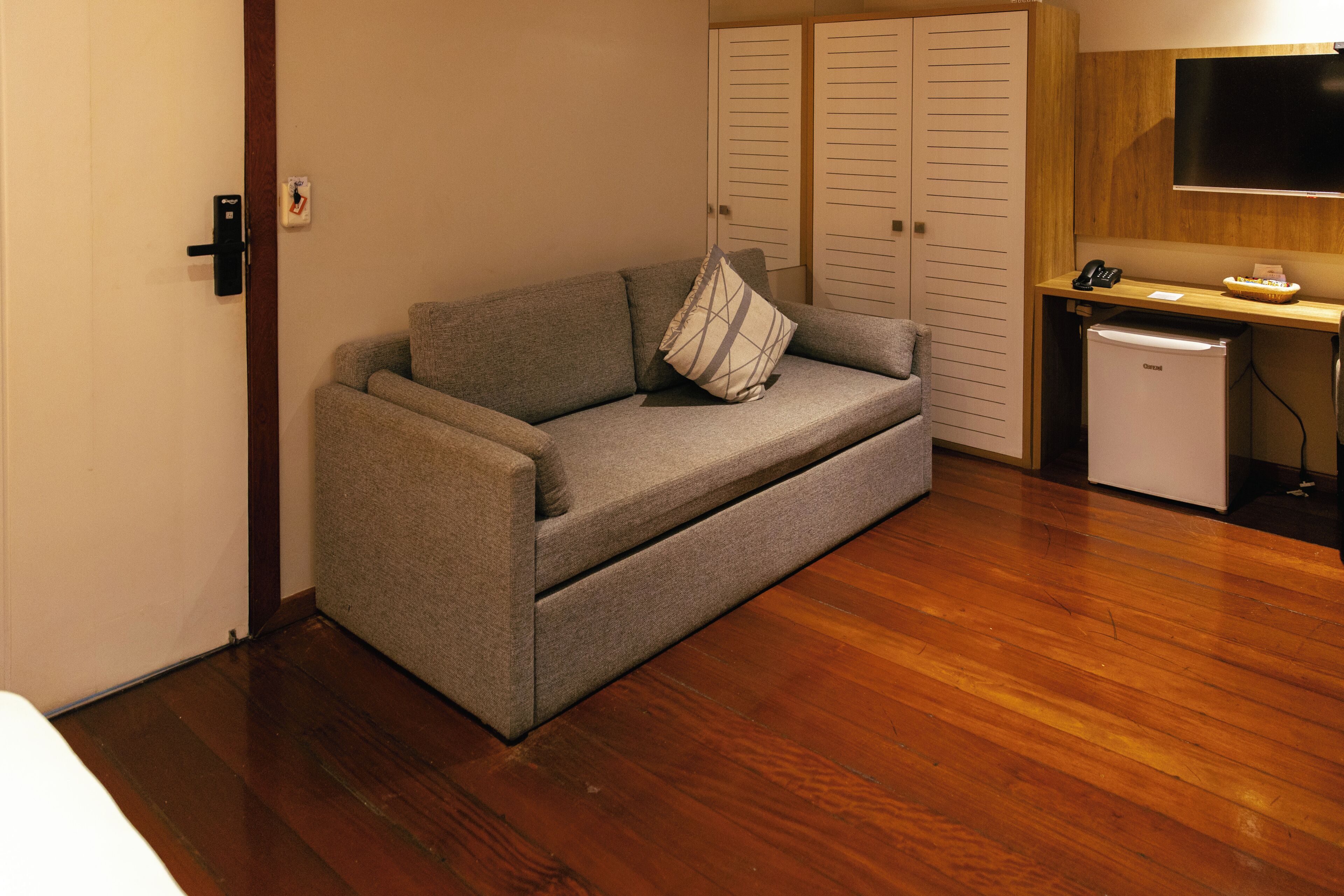 A compact room with a grey sofa, a small wooden desk, a white refrigerator, and a wardrobe with multiple doors.