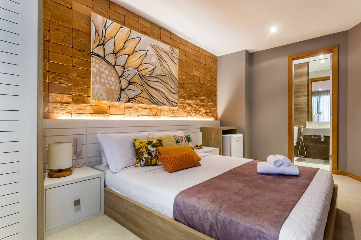 Une chambre moderne avec un grand lit, une table de chevet, une commode, une œuvre d’art accrochée au mur et une salle de bain visible à travers une porte ouverte.