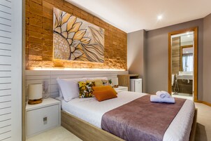 Un dormitorio moderno con una cama grande, una mesita de noche, una lámpara auxiliar y una obra de arte en la pared, con un baño visible a través de una puerta abierta.