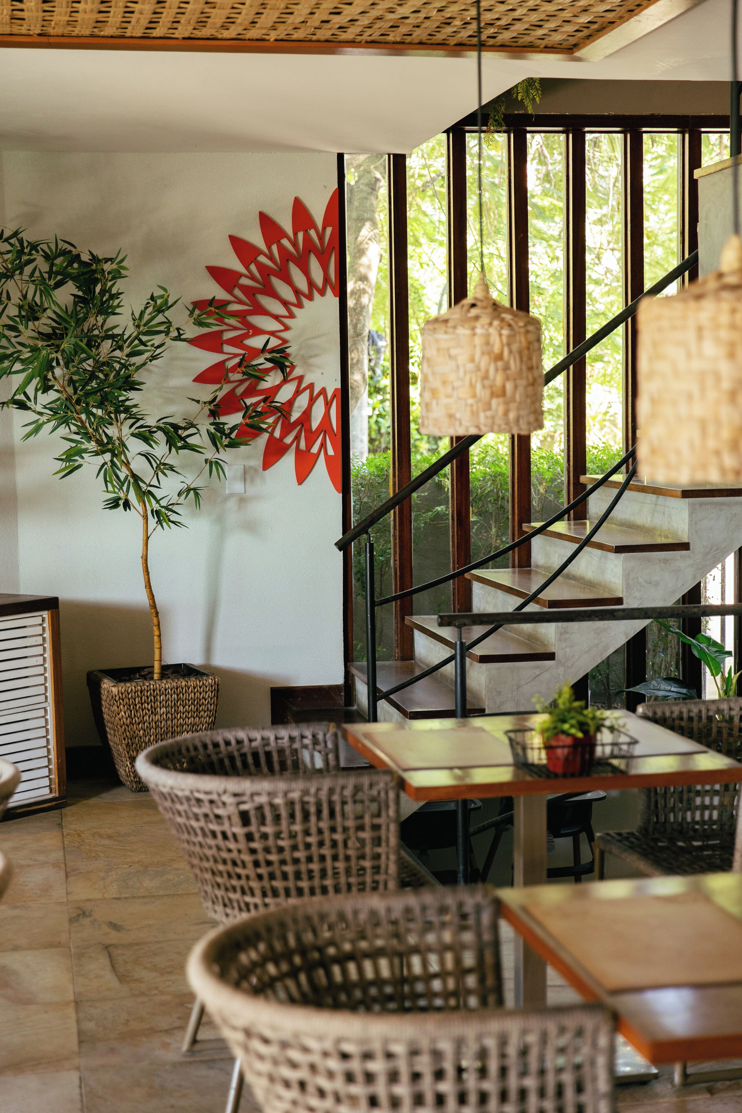 A restaurant interior with wicker furniture, a staircase, and a large window with a view of greenery.
