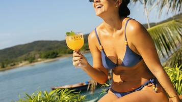 Eine Person in einem blauen Bikini sitzt in einem Whirlpool mit einem Getränk, umgeben von Grünpflanzen und einem Gewässer.