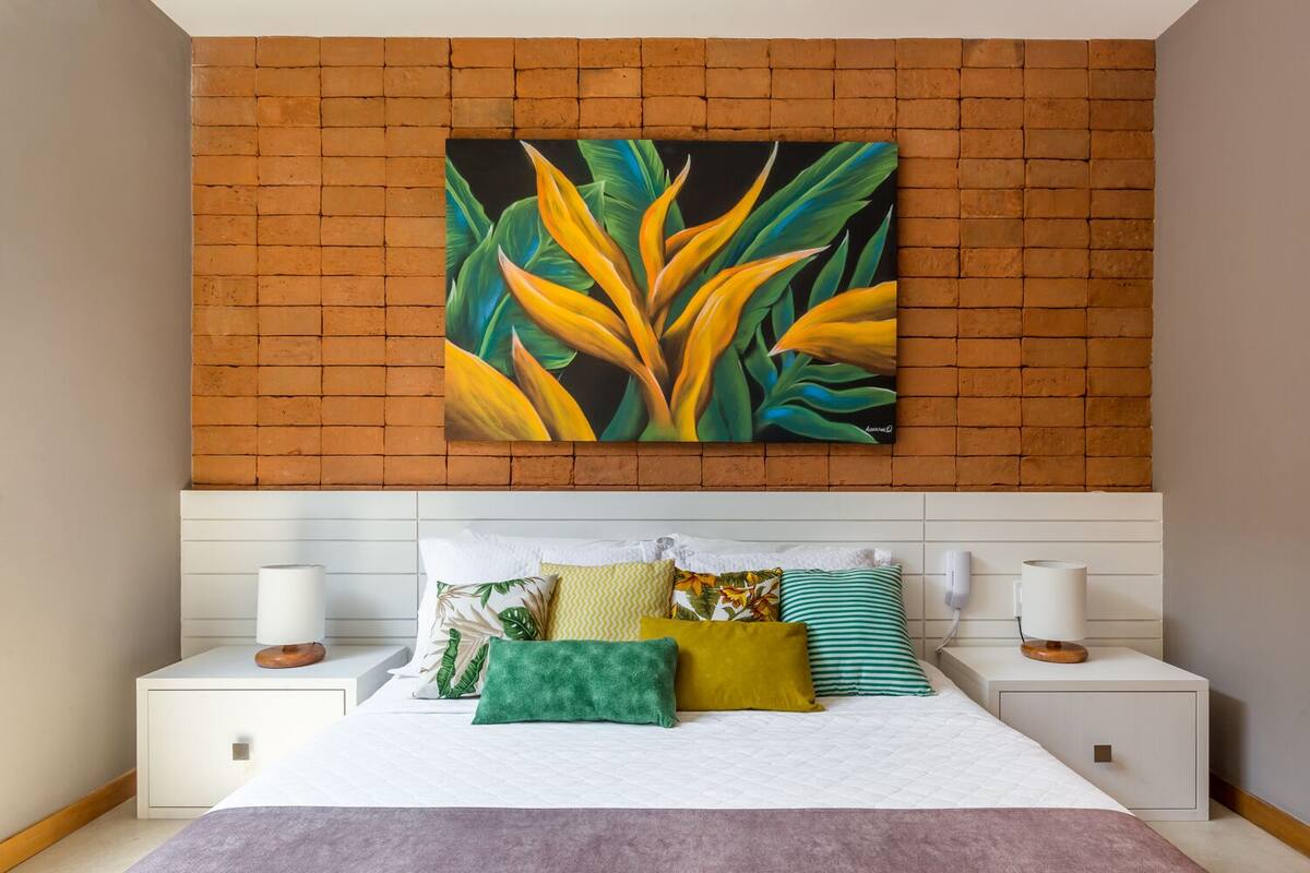 Une chambre avec un mur en briques, un lit recouvert de plusieurs coussins et un tableau représentant des feuilles tropicales.