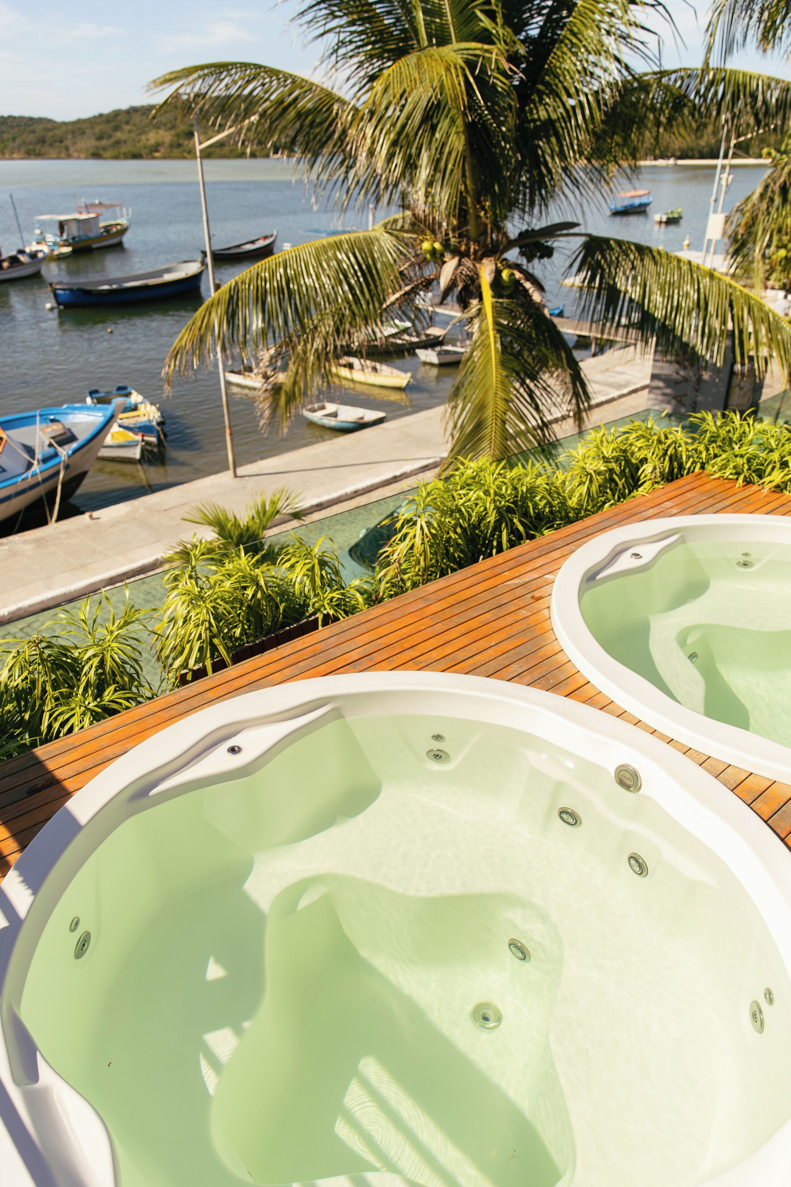 Área à beira da piscina com duas banheiras de hidromassagem, um deck de madeira e vista para barcos na água.