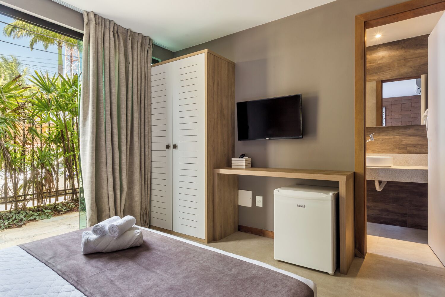 A hotel room with a bed, a TV, a mini-fridge, a bathroom with a sink, and a view of palm trees.