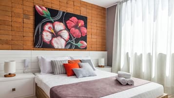 Una habitación de hotel moderna con una pared de ladrillo decorativa, un cuadro floral grande, una cama bien hecha con varias almohadas, una mesita de noche con lámpara y una ventana con cortinas de gasa.
