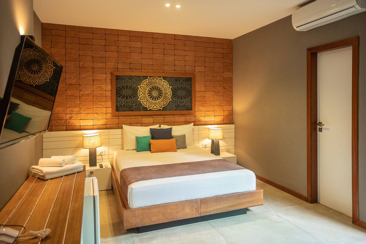 Une chambre d’hôtel moderne avec un mur d’accent en briques, un grand lit avec de nombreux oreillers, des lampes de chevet, une table de chevet en bois avec un téléphone et une porte.