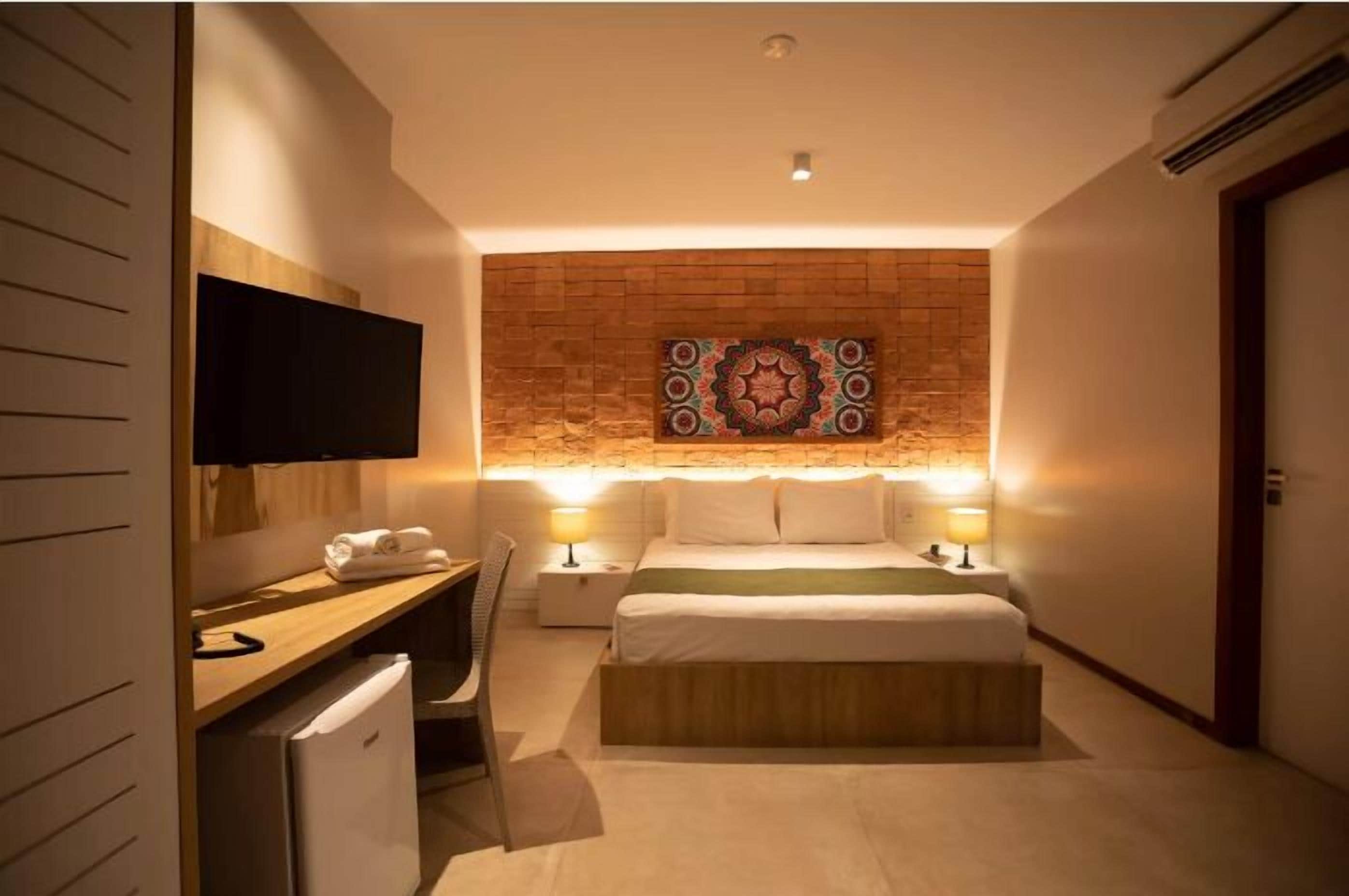 Habitación de hotel con cama, lámparas de noche, televisor de pantalla plana, escritorio con silla y un refrigerador pequeño.