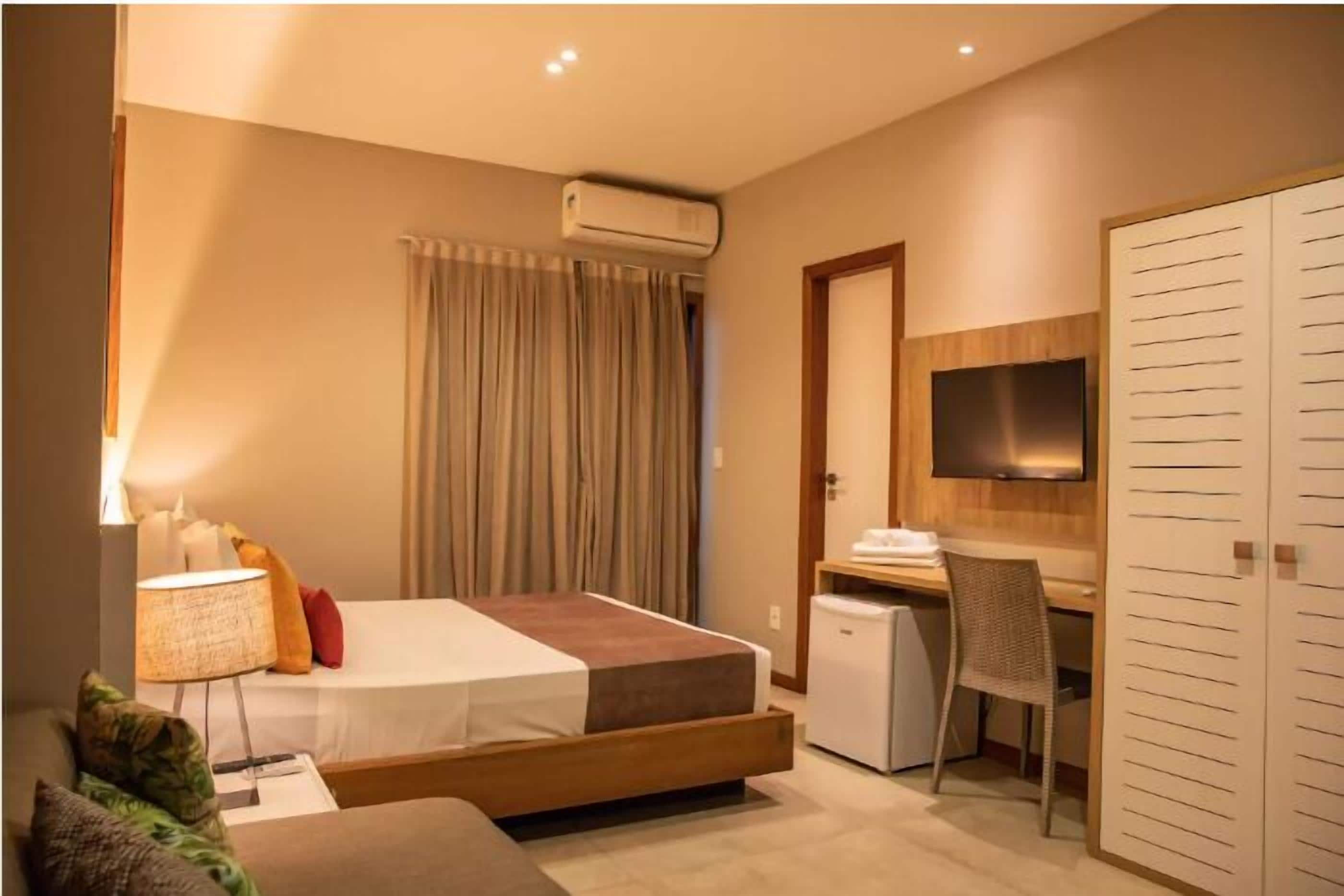 Quarto de hotel com cama, escrivaninha, cadeira, televisão, guarda-roupa e ar condicionado.