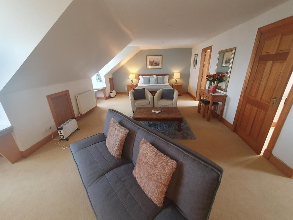 A loft-style bedroom with a sloped ceiling, a large bed, two bedside tables with lamps, a sofa, and a dining table.