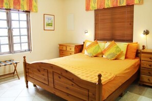 A wooden bed with a yellow bedspread and leaf-patterned pillows, a wooden nightstand, a wooden dresser, and a window with a view of palm trees.