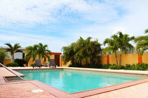 A swimming pool with tiled edges, surrounded by palm trees and a yellow wall.