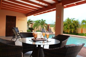 A covered outdoor seating area with wicker chairs and a round table, overlooking a swimming pool and palm trees. - Sunrise Villa (Noord)