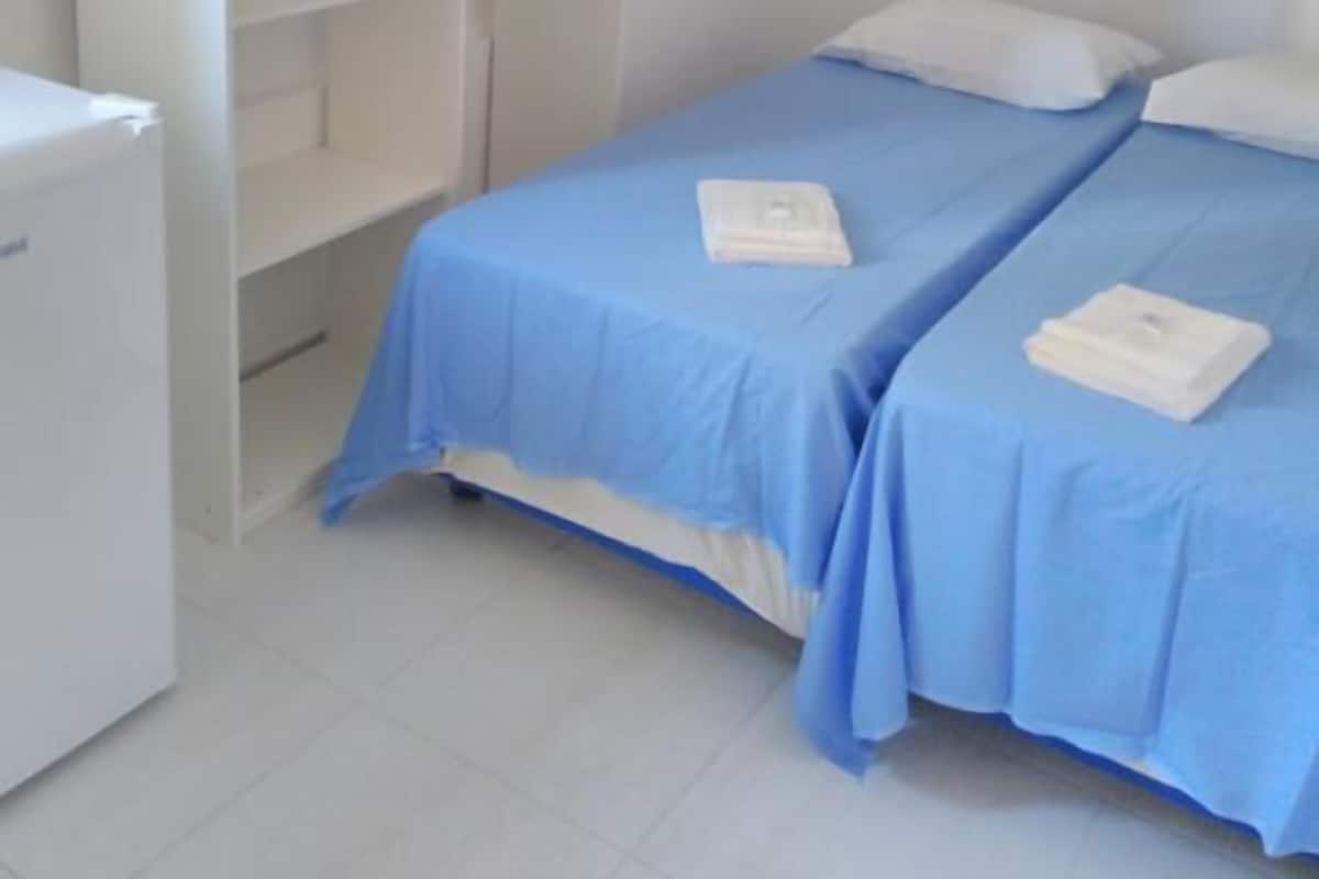 A room with two beds, each with a blue bedspread and white pillows, and a white wardrobe with open doors.