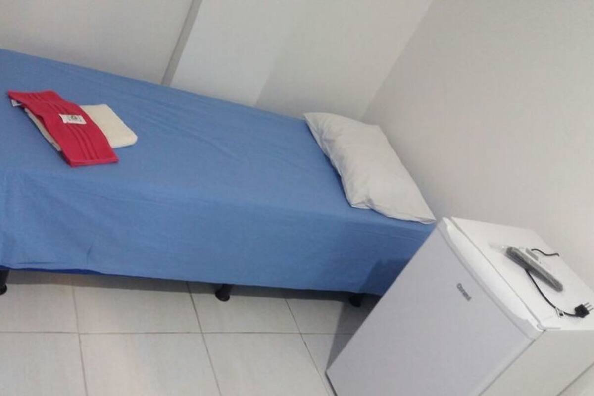 A small, single-bed room with a blue bedspread, a white pillow, and a red item on the bed. There is a white cabinet with a power strip on top.
