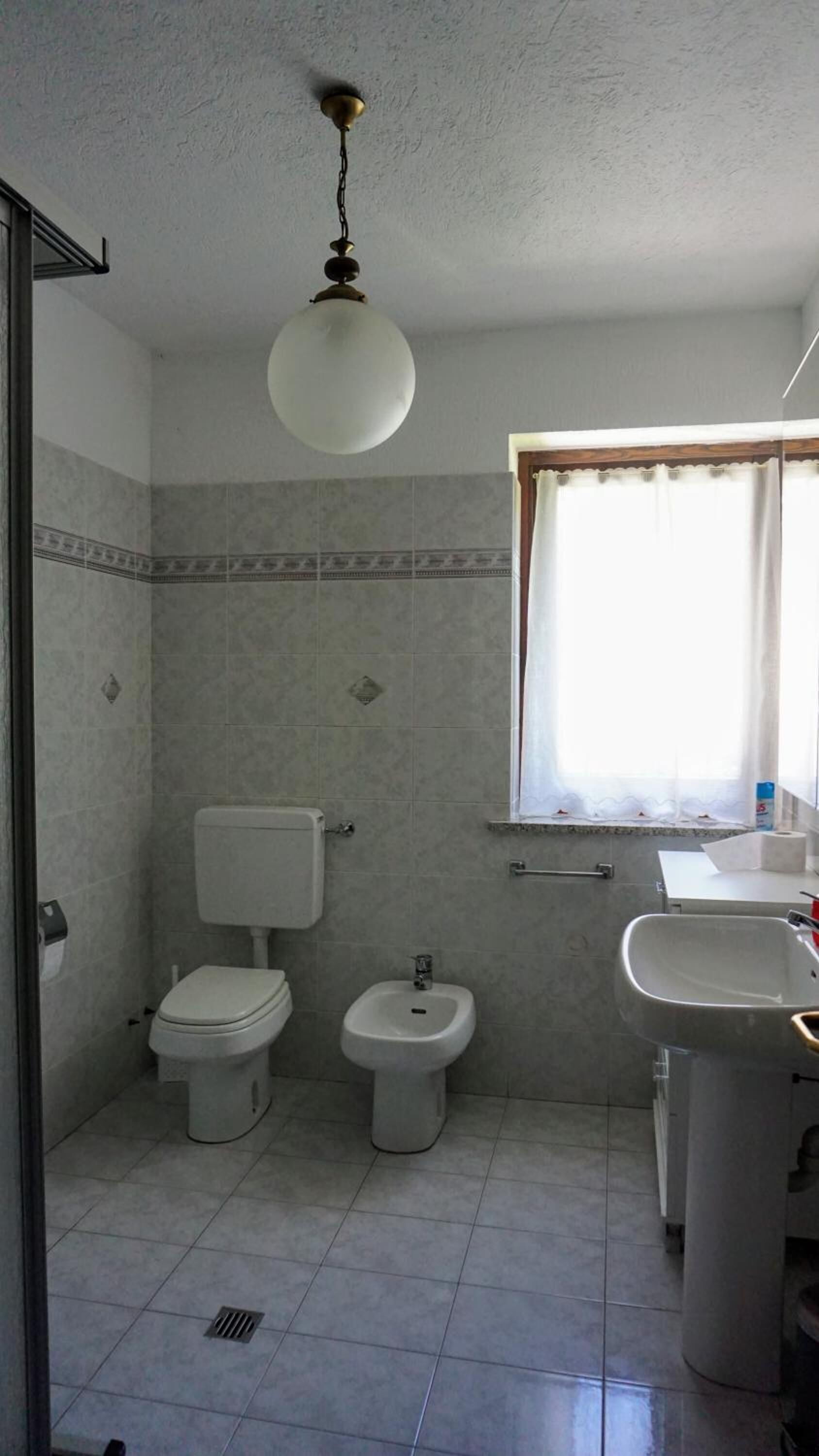 Un baño con inodoro, bidé y lavamanos, paredes con azulejos y una ventana con cortinas.