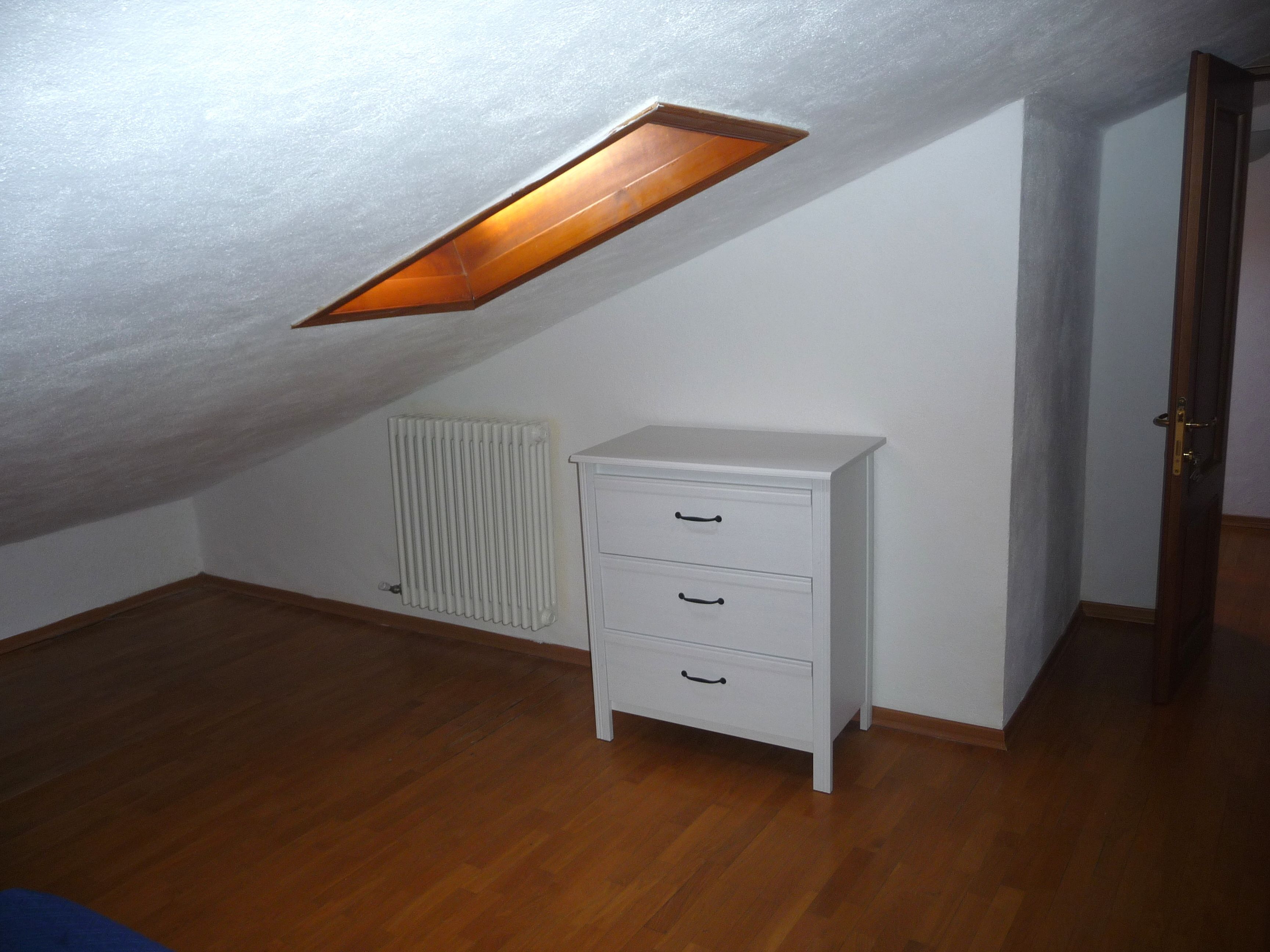 A room with a sloped ceiling, a radiator, a white dresser with drawers, and a door.