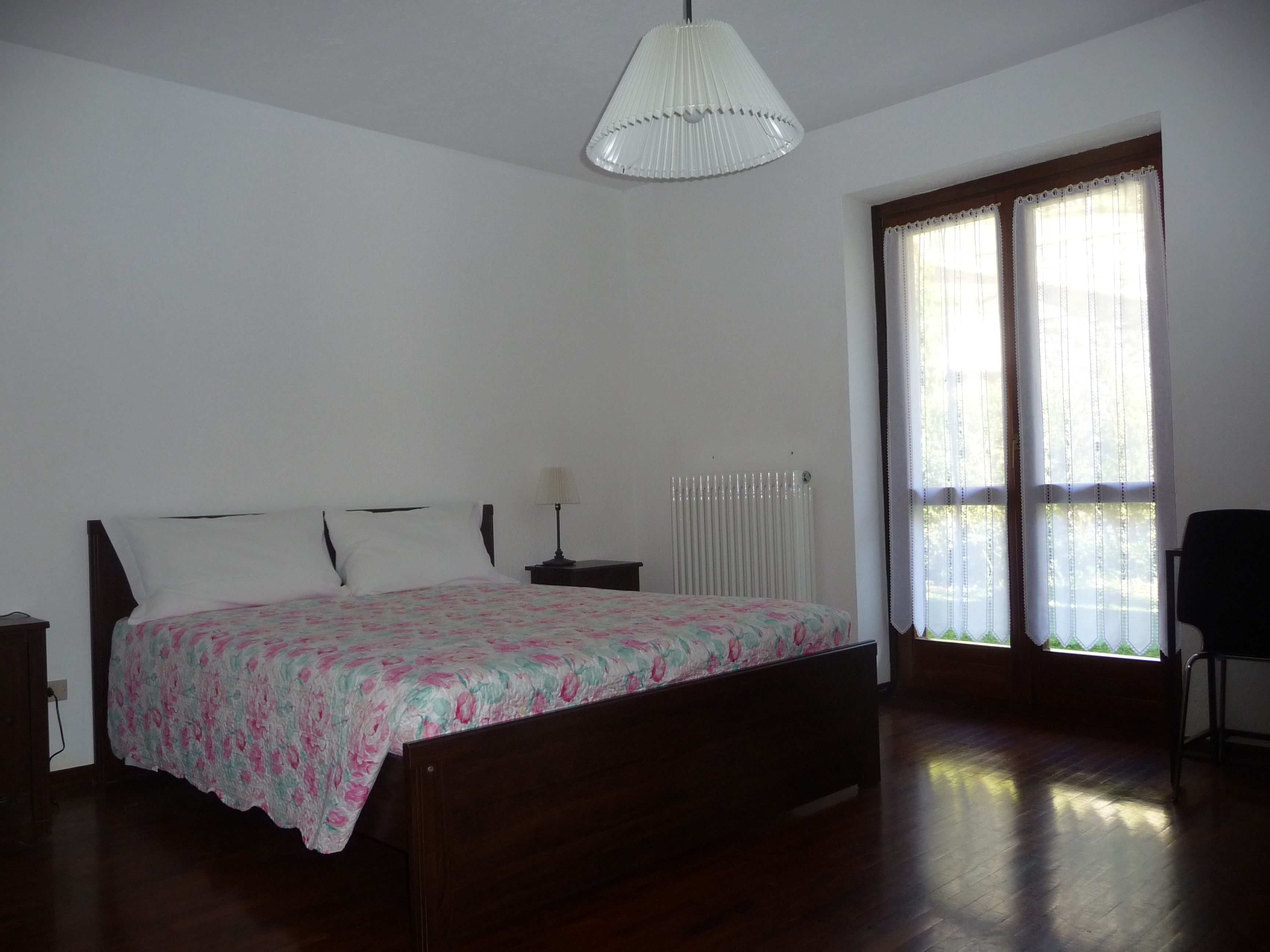 Ein Schlafzimmer mit einem hölzernen Bett, einem Nachttisch, einer Lampe, einer Heizung und einem Fenster mit Vorhängen.
