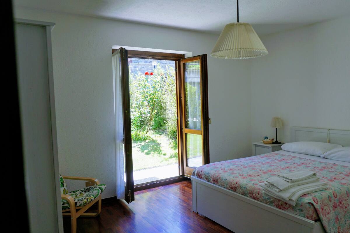 A bedroom with a bed, a nightstand, a lamp, a chair, and a window with a view of greenery.