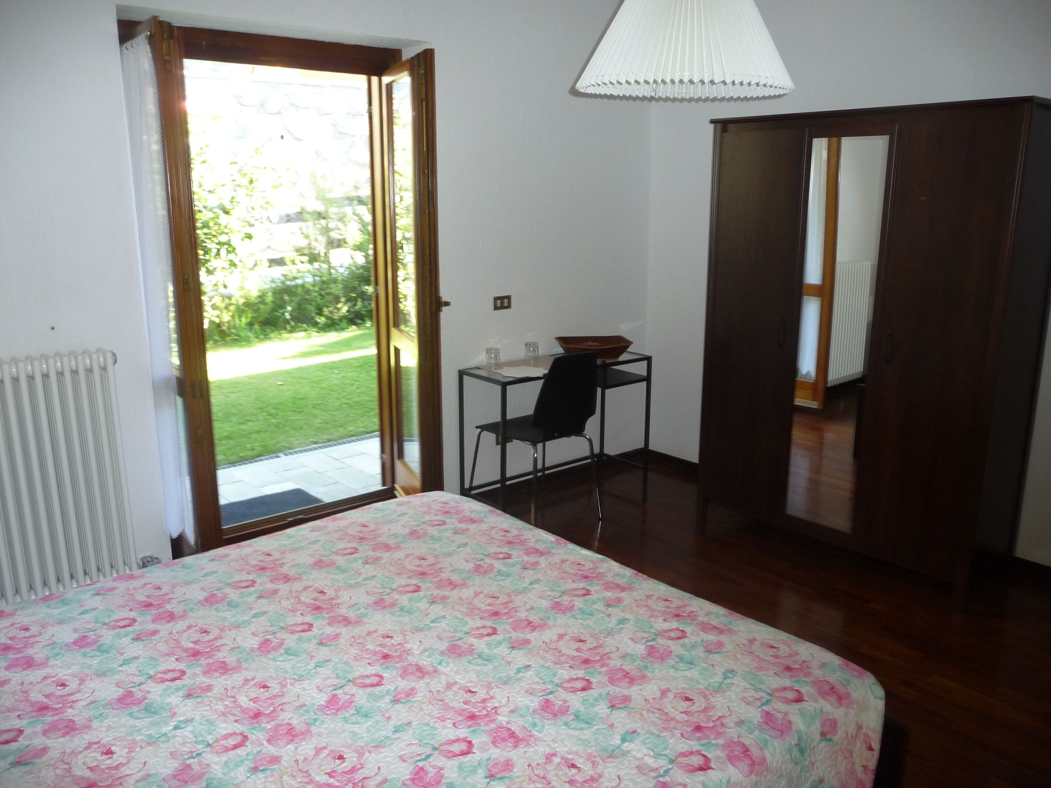 Ein Schlafzimmer mit Bett, einem kleinen Tisch mit Stuhl, einem Heizkörper und einer Tür, die auf einen Balkon führt.
