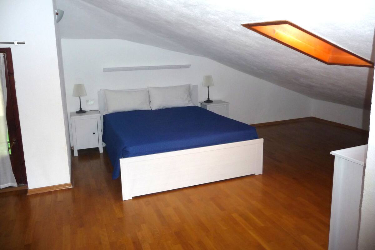 A bedroom with a sloped ceiling, a bed with a blue bedspread, two bedside tables with lamps, and a wooden floor.