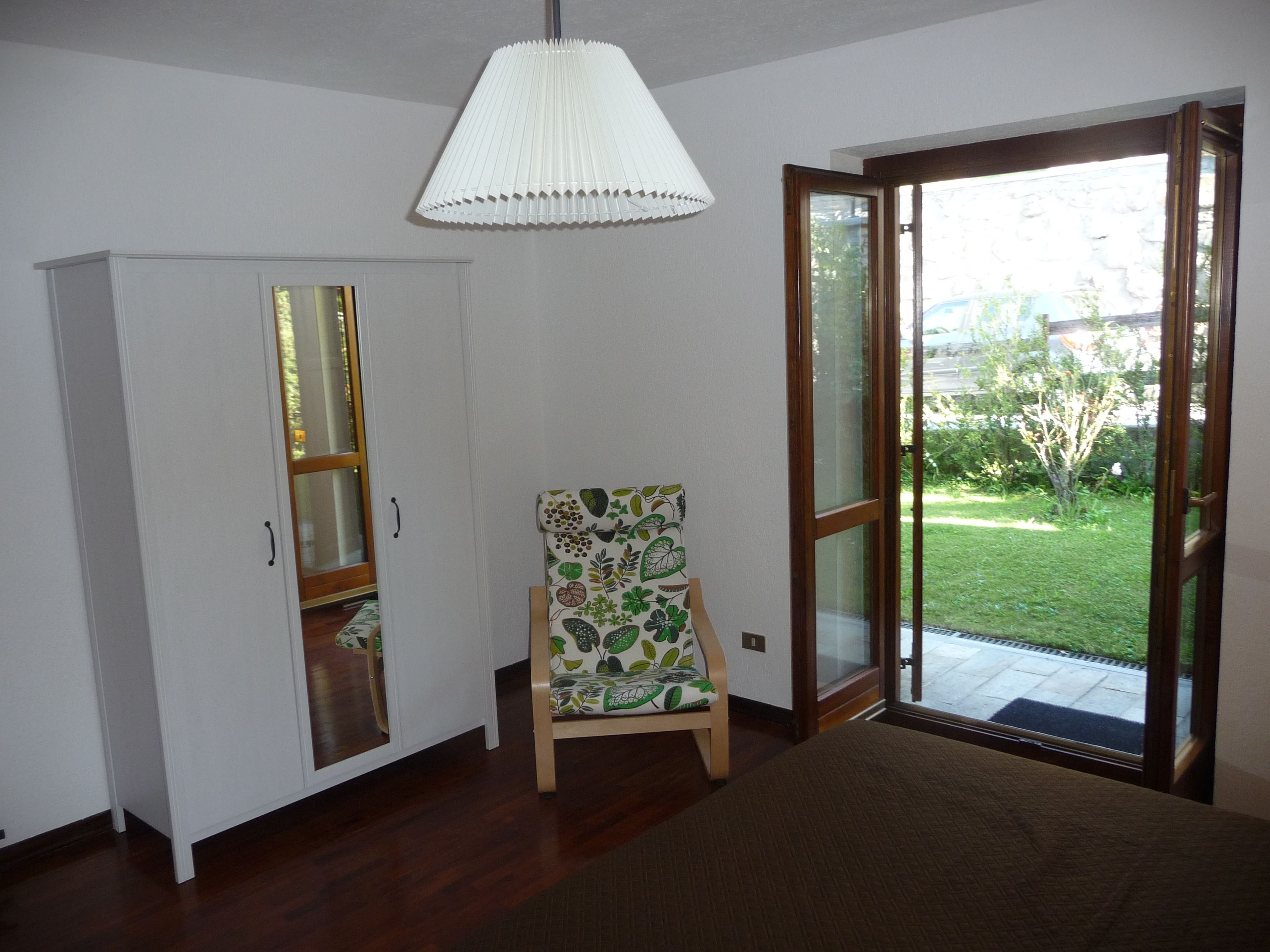 A room with a wooden floor, a white wardrobe, a patterned armchair, and a sliding glass door leading to a garden.