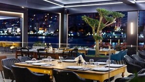 A restaurant with tables set for dining, a large window with a view of the city at night, and a tree in the center. - Demi Hotel (Sarandë)
