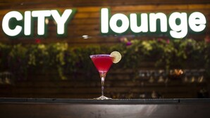 Un verre à martini, agrémenté d’une garniture, posé sur le comptoir d’un bar, avec une enseigne « CITY lounge » en arrière-plan.