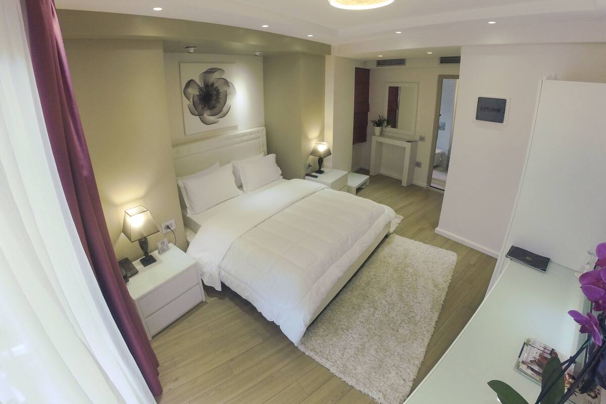 A modern bedroom with a large bed, bedside lamps, a ceiling light, a mirror, and a small table with a plant.