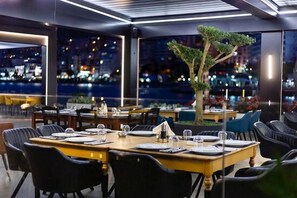 Un restaurant avec des tables dressées pour le dîner, une grande fenêtre offrant une vue sur la ville la nuit, et un arbre au centre.