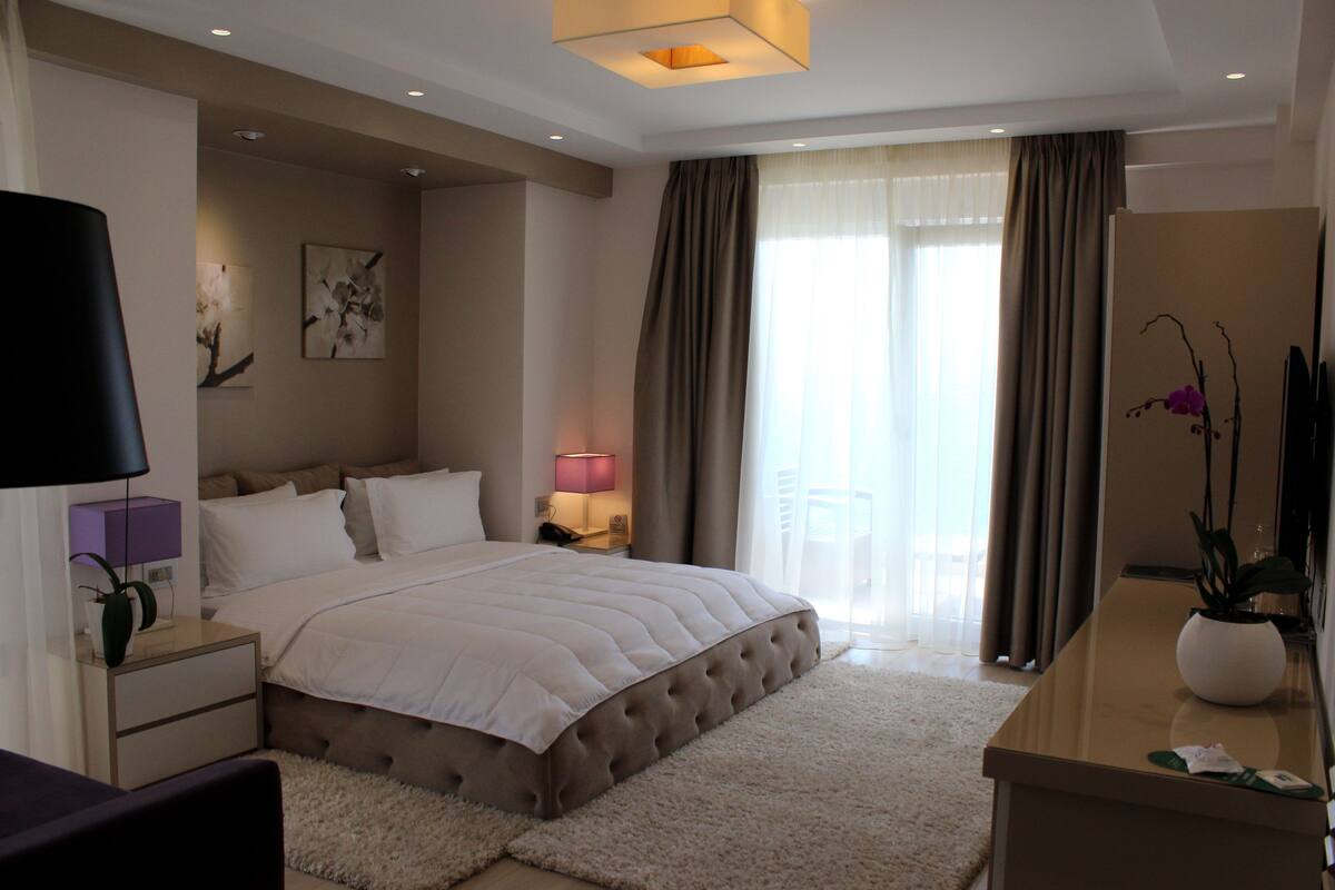 A modern hotel bedroom with a large bed, a bedside table with a lamp, a nightstand, a desk with a vase, and a window with curtains.