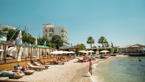 A beach with sun loungers and umbrellas, a building labeled "DEMI HOTEL" in the background, and a clear blue sky. - Demi Hotel (Sarandë)